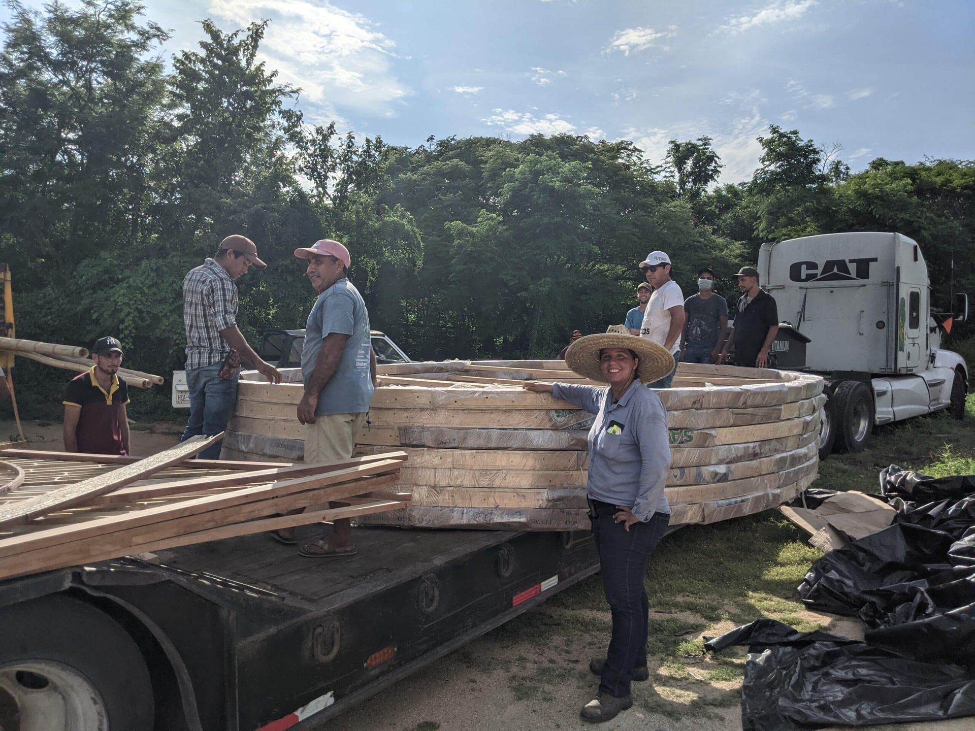 Workers load stacked lumber onto a flatbed trailer beside a white truck outdoors.
