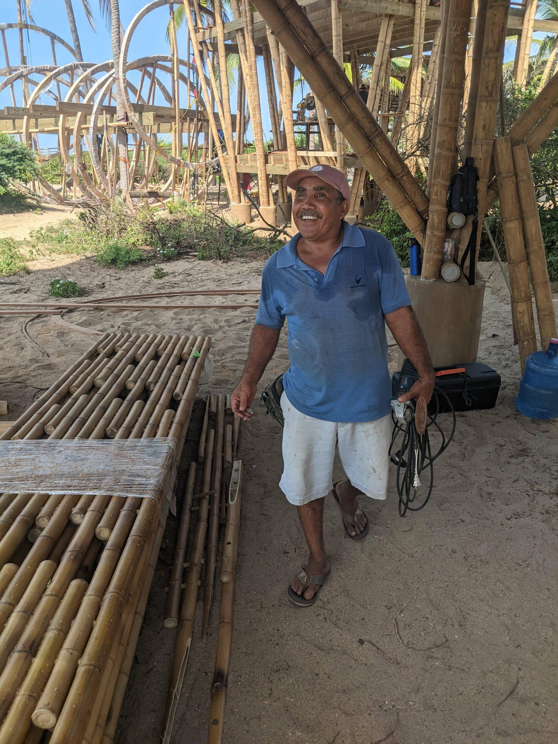 Smiling person in a blue shirt and white shorts stands beside bamboo structures outdoors.