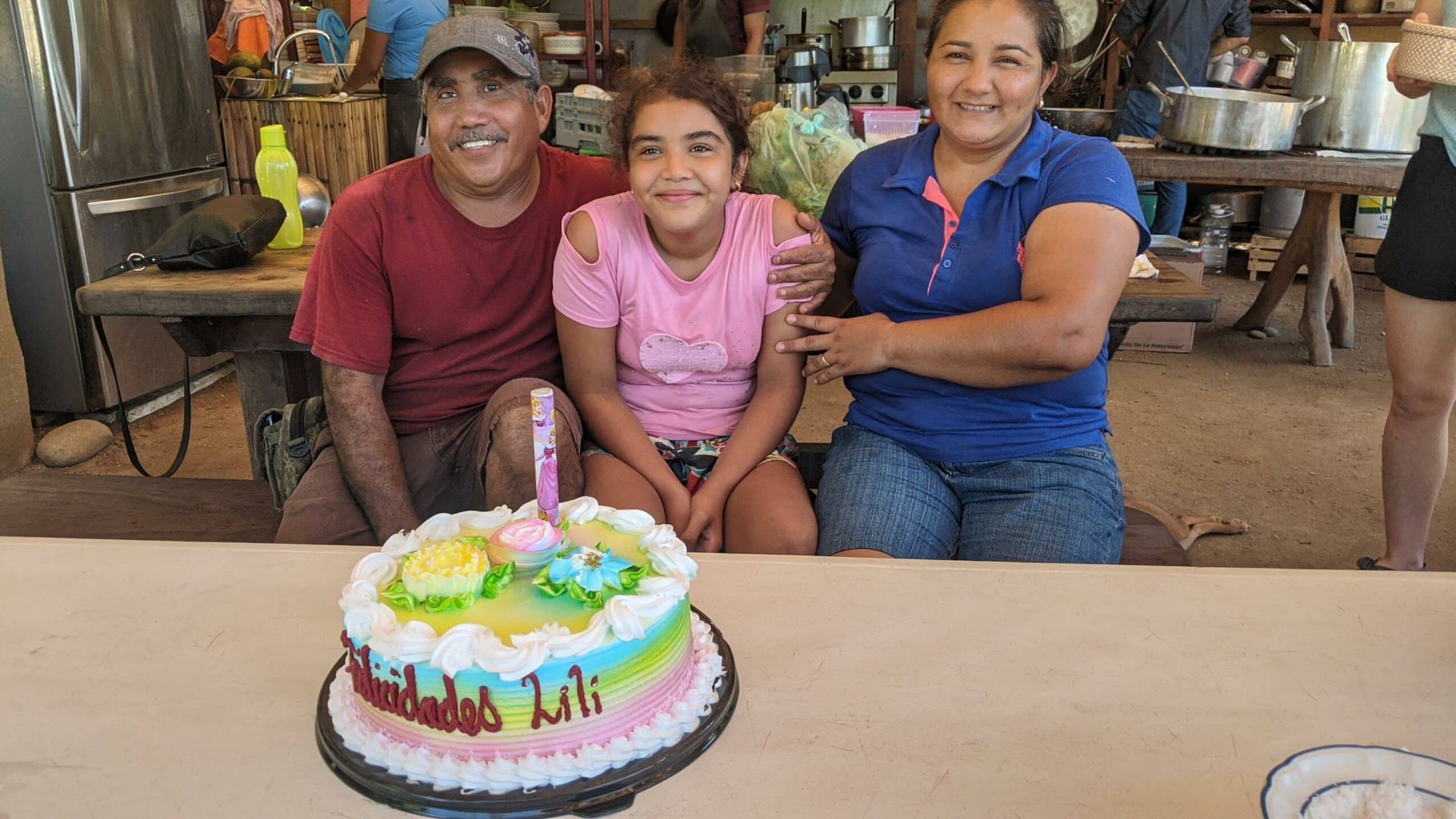 Three people smiling behind a colorful birthday cake at a table indoors
