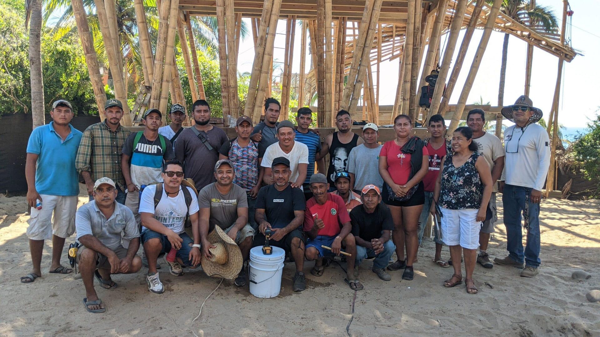 Group of people posing outdoors under a wooden shelter on a sandy beach, with buckets in front.
