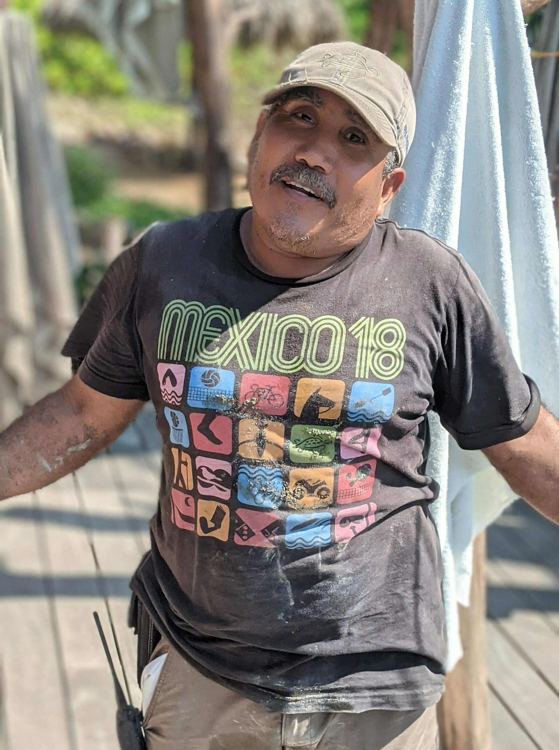 Person in a beige cap and black graphic T-shirt standing outdoors by a hammock, smiling.