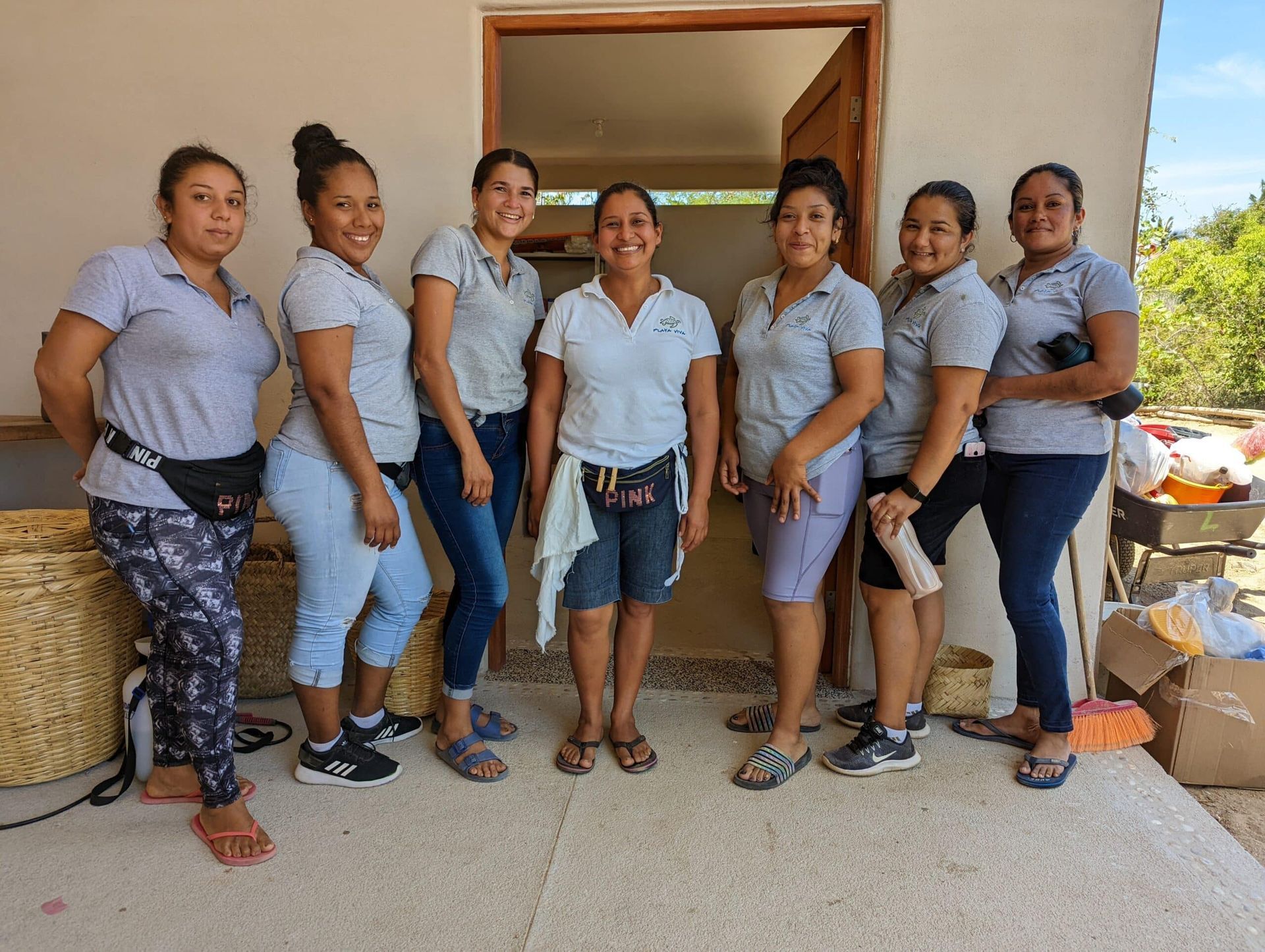 Seven people standing together in a doorway, wearing light blue shirts and patterned leggings, smiling outdoors
