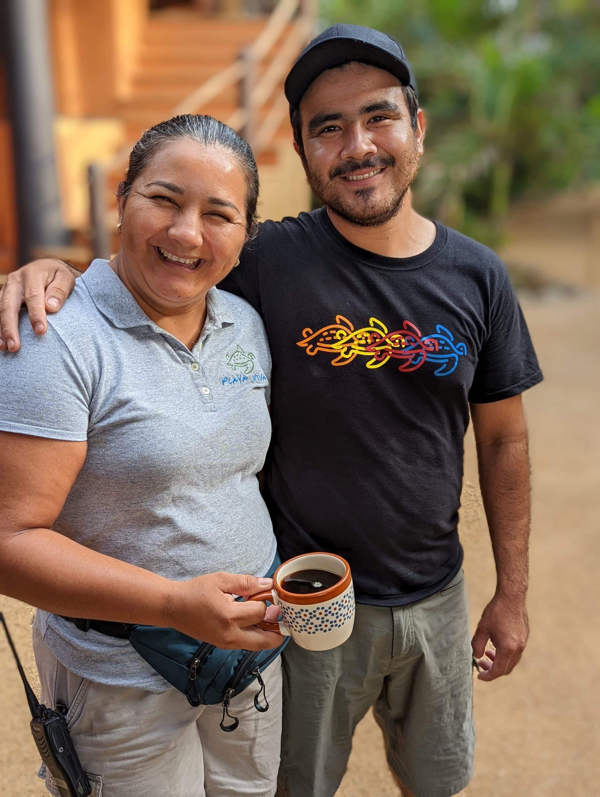 Two people standing outdoors, smiling, with one holding a mug and the other’s arm around them.