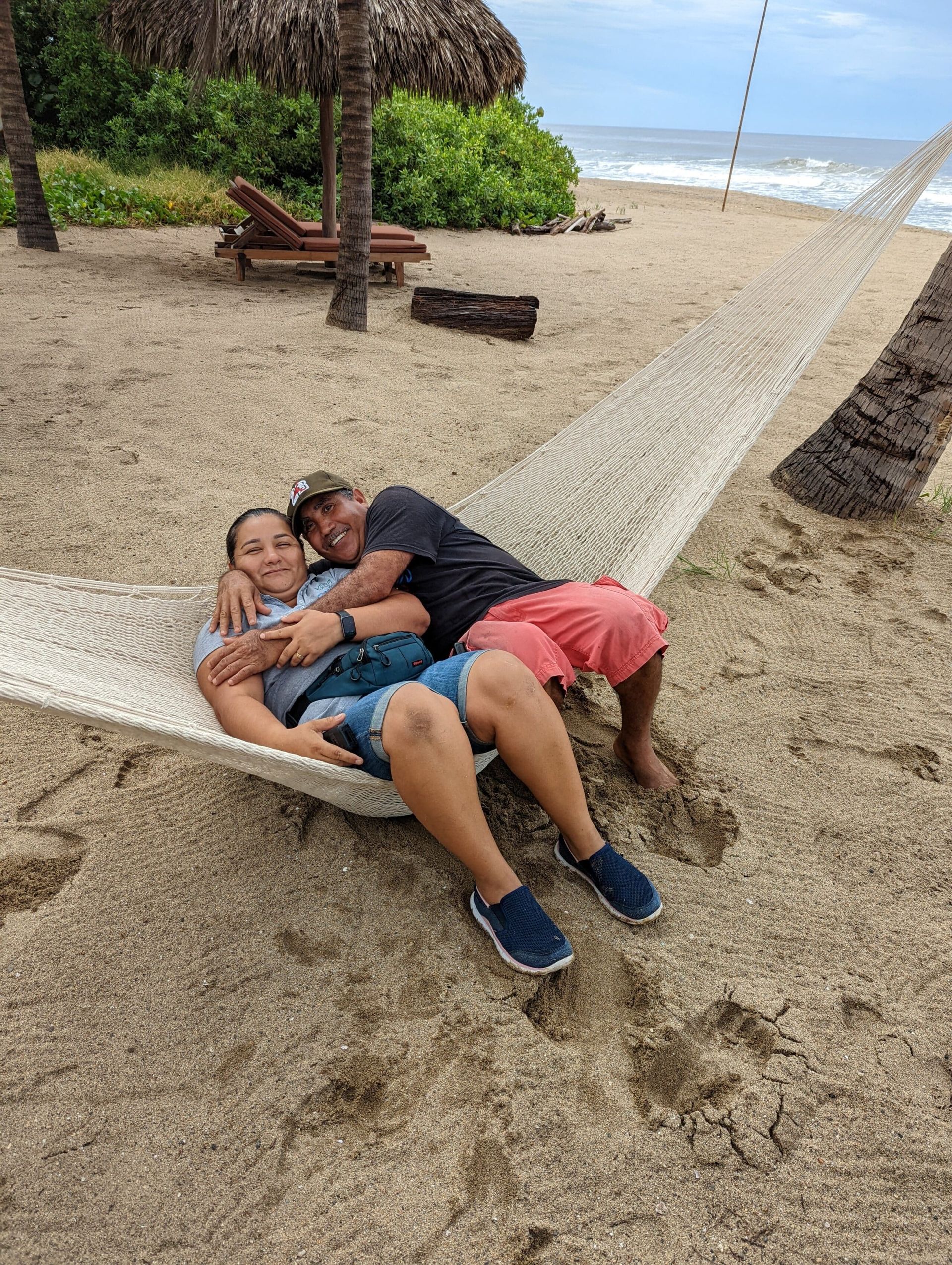 Two people relaxing together on a beach hammock near the shore, with palm trees and ocean in the background.