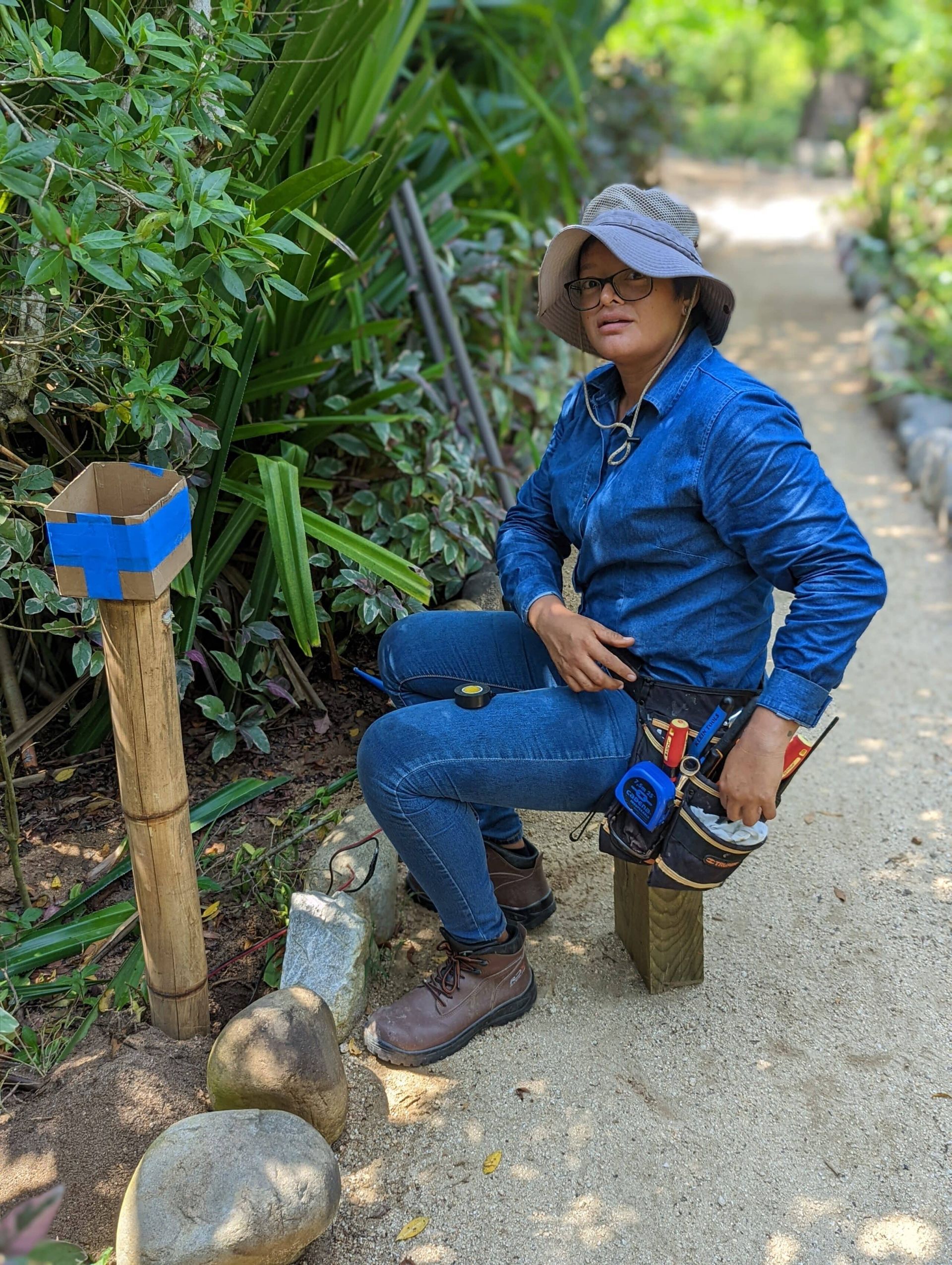 Person in blue work clothes sitting on a low stool beside a garden path and plants, holding tools.
