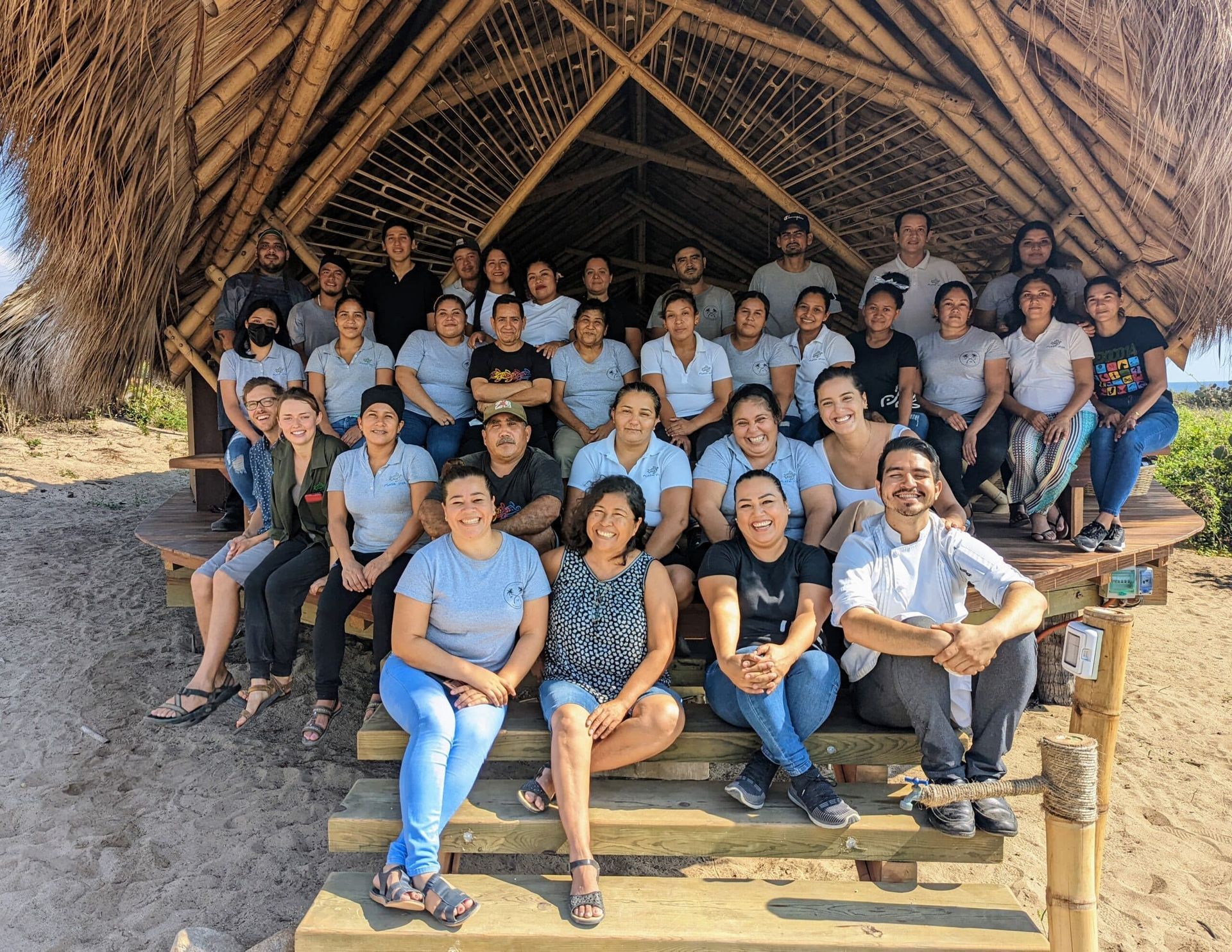 Group of people posing on wooden steps beneath a thatched shelter, many wearing light blue shirts.