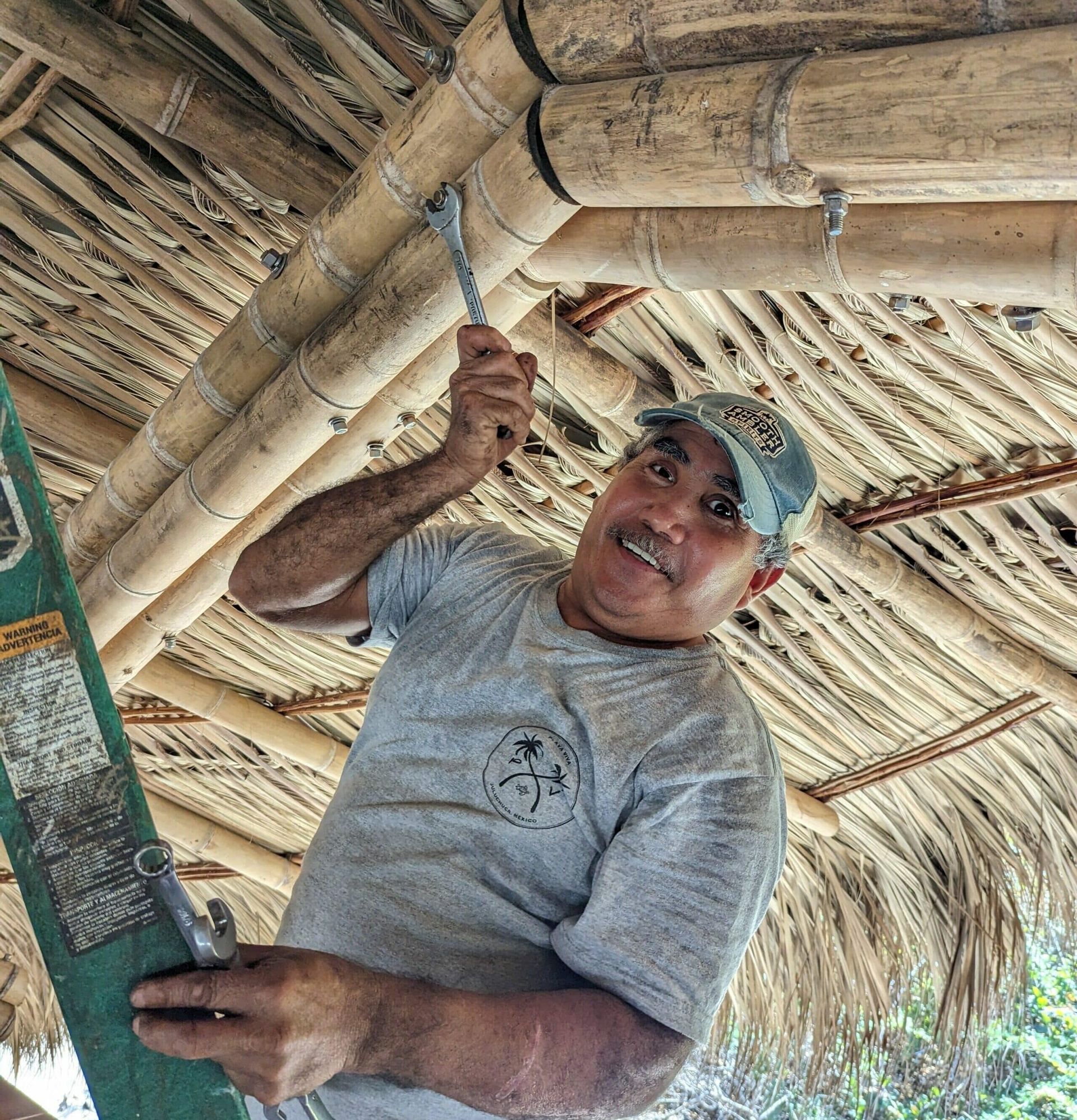 Man smiling while working overhead in a thatched-roof structure, holding a tool and a green board