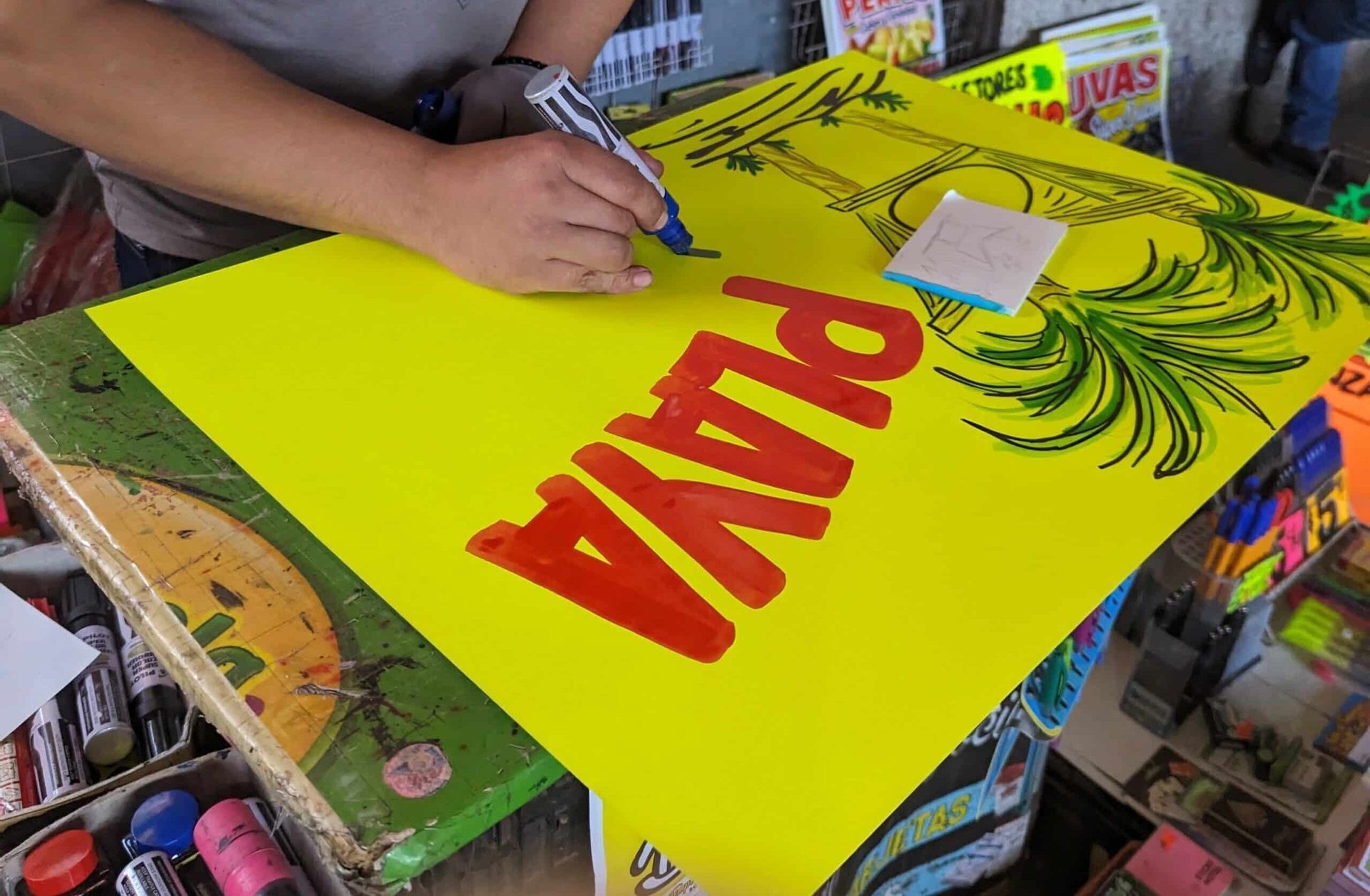 Hands painting a bright yellow protest sign with large red letters and a green leaf design