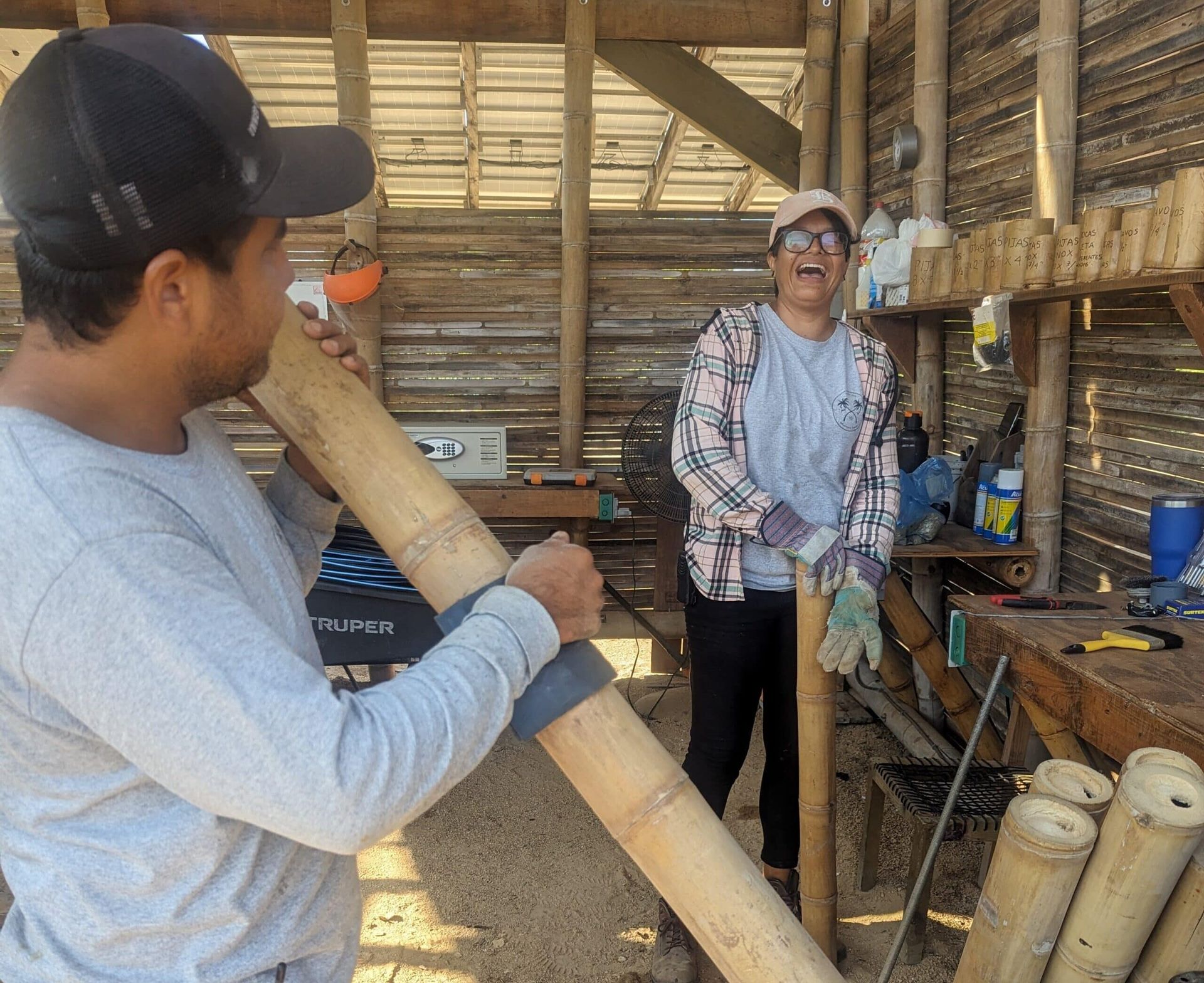 Two people in a rustic workshop handling a long bamboo pole and smiling near tools and stacked bamboo pieces.
