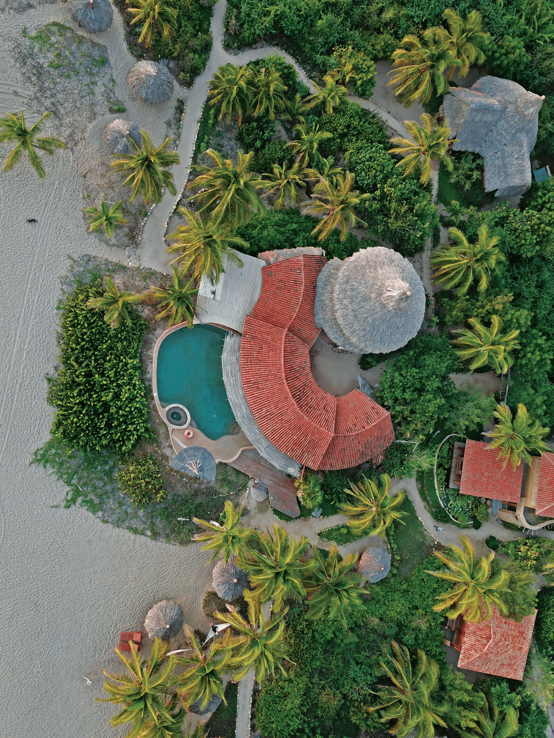 Aerial view of a tropical resort with a curving pool, thatched-roof huts, palm trees, and sandy paths