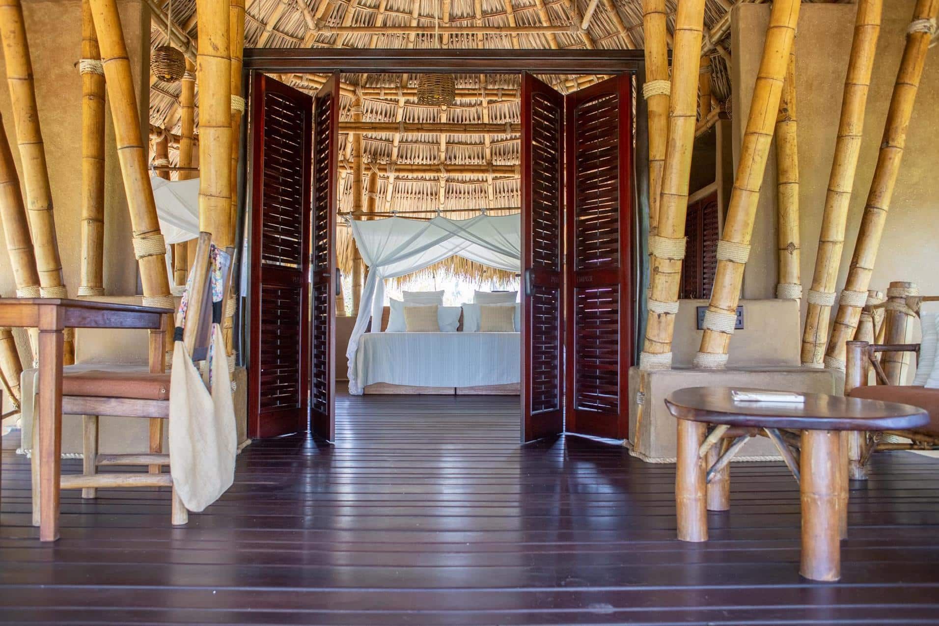 Bamboo lodge interior with open double doors, wooden tables, and a bed draped in white beyond the doorway