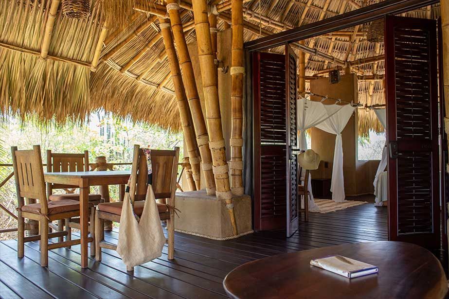 A wooden table and chairs are sitting on a wooden deck under a thatched roof.