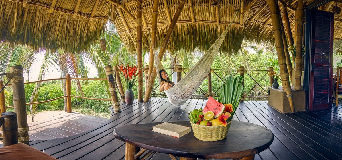 Open-air thatched hut with hammock, round table, and a fruit basket overlooking lush greenery