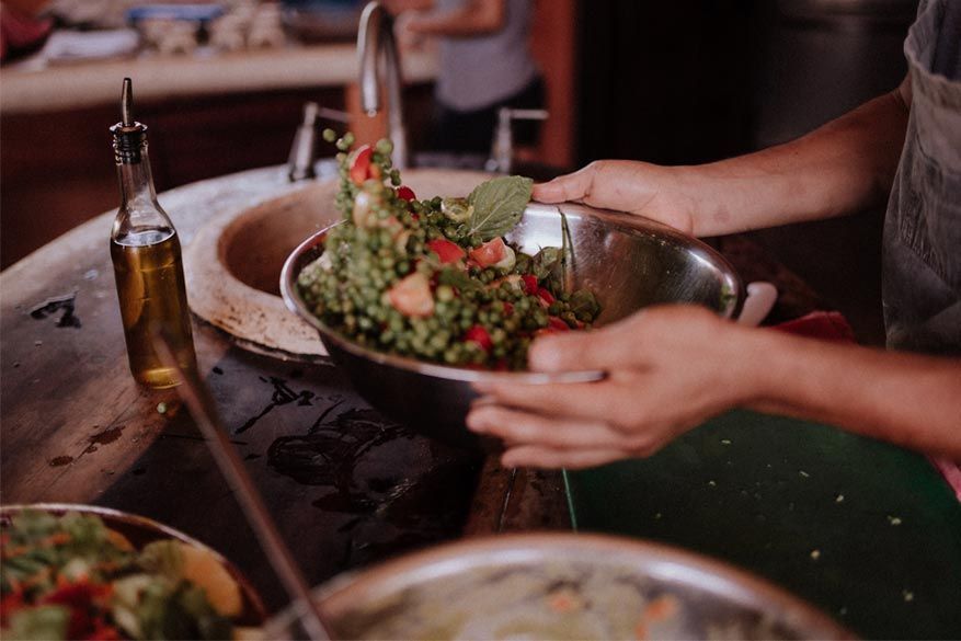 Hands serving a salad in a bowl beside a bottle of olive oil on a dining table