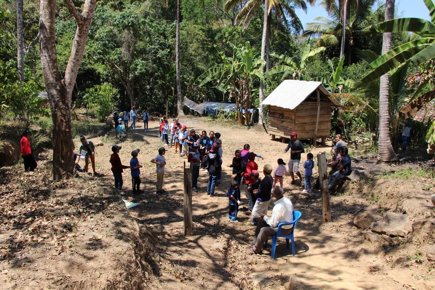 People gathered outdoors in a rural clearing around a small structure, with trees and dirt ground.