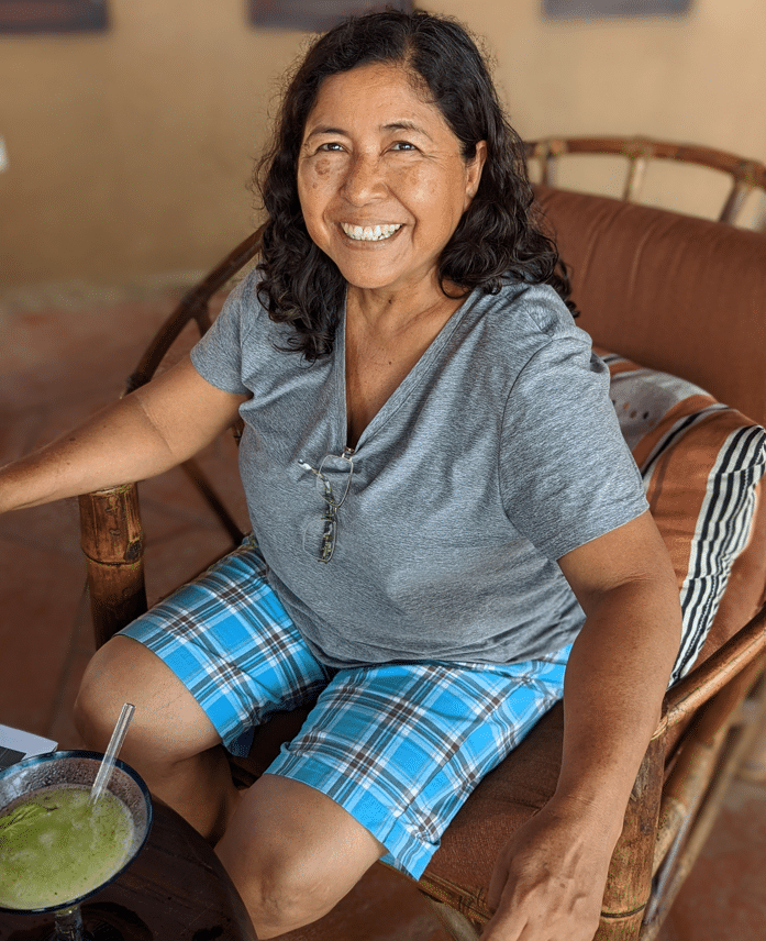 Smiling woman in a gray shirt and blue plaid shorts sitting on a wicker chair, holding a coconut drink.