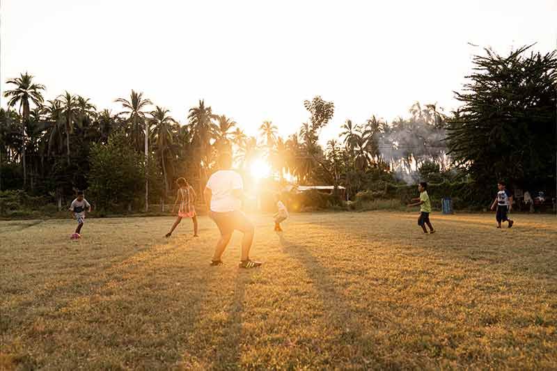 People playing soccer on a grassy field at sunset, with the sun low on the horizon.