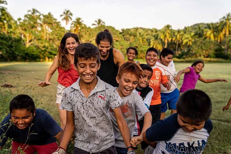 Children laughing and running together on a grassy field with palm trees in the background.