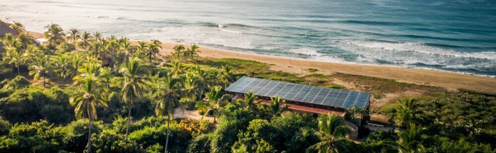 Aerial view of a coastal tropical resort with palm trees, solar panels, and ocean waves beyond.