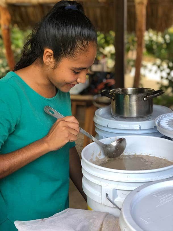 Woman stirring food in large white buckets beside a pot, smiling in an outdoor kitchen.