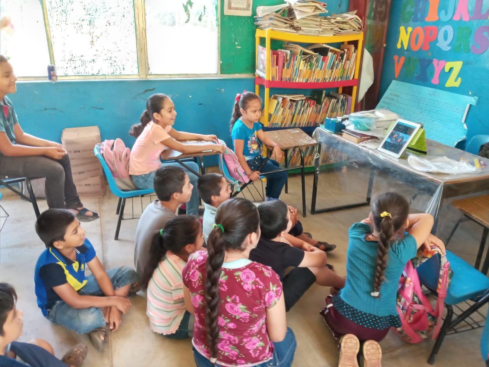 Children sitting in a classroom, watching a lesson near a table with books and a screen