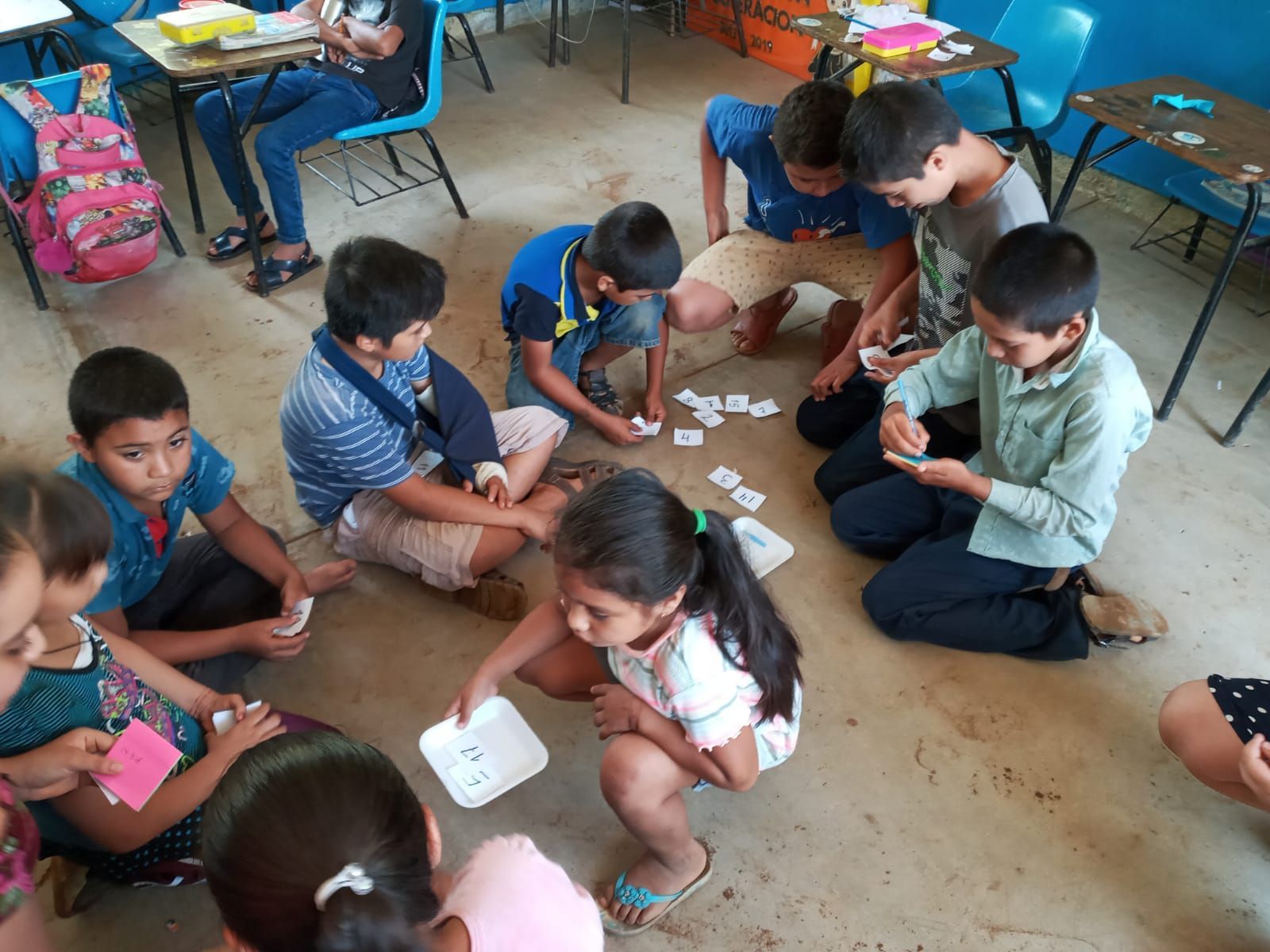 Children sitting on the floor in a classroom, playing a card game together with scattered cards.
