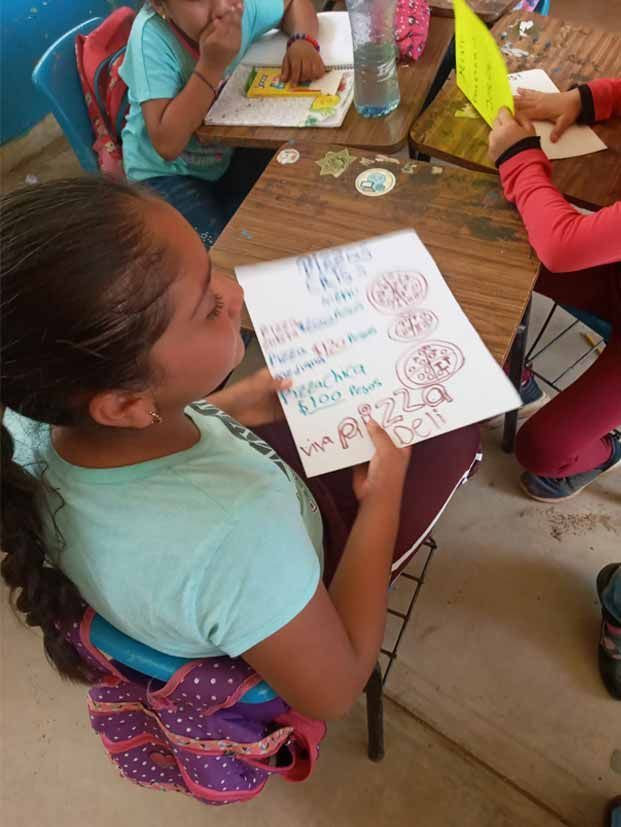 Child seated at a table, holding a worksheet and writing with colorful materials in a classroom.