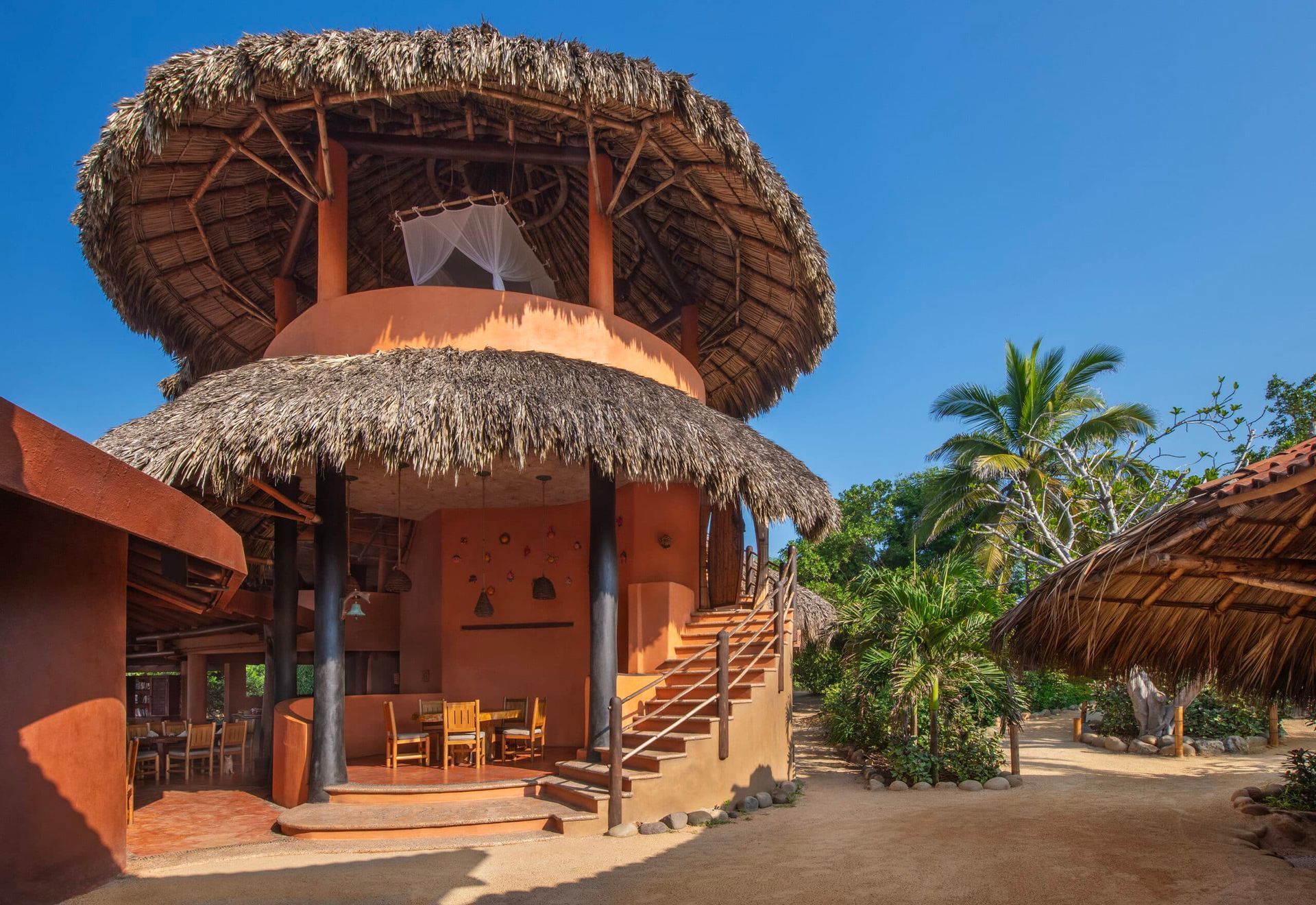 Thatched tropical lodge with a round upper level, palm trees, and a sunny outdoor patio