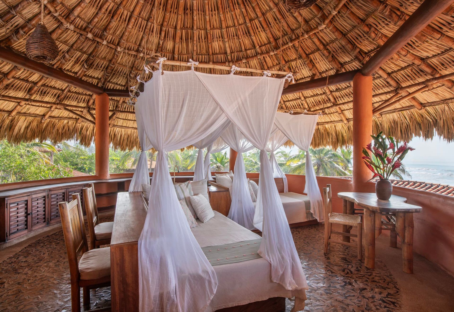 Thatched beachfront cabana with a four-poster bed draped in white netting and a small table overlooking greenery
