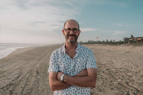 Man standing on a sandy beach with arms crossed, smiling at the camera under a cloudy sky
