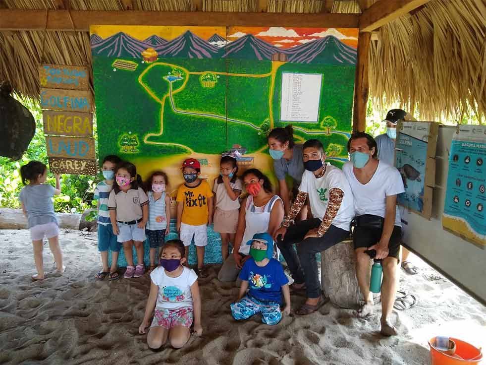 Group of masked people in a rustic classroom, posing before a green chalkboard with a yellow drawing.