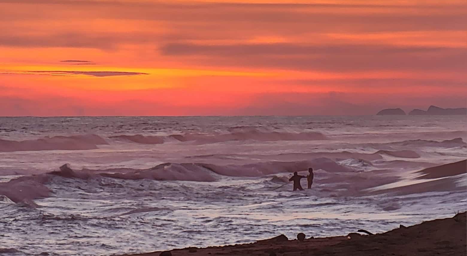 Two people walking on a rocky beach under a pink-orange sunset with rough waves.
