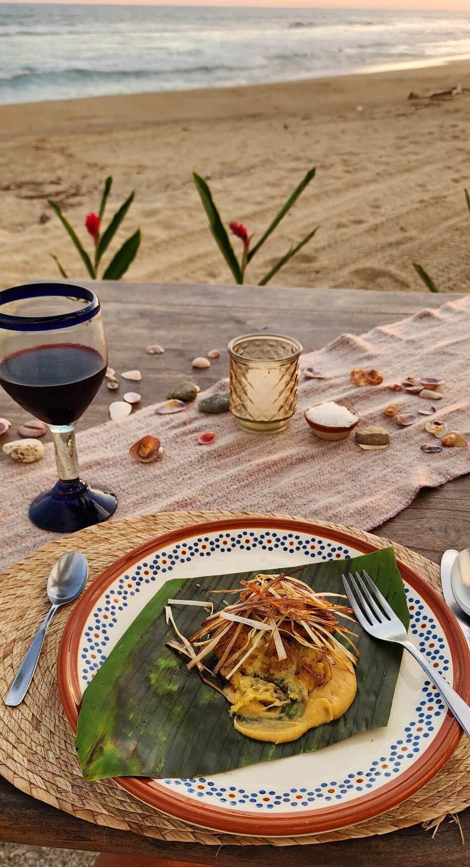 Beachside dinner table with wine, candle, and plated meal on a patterned cloth at sunset