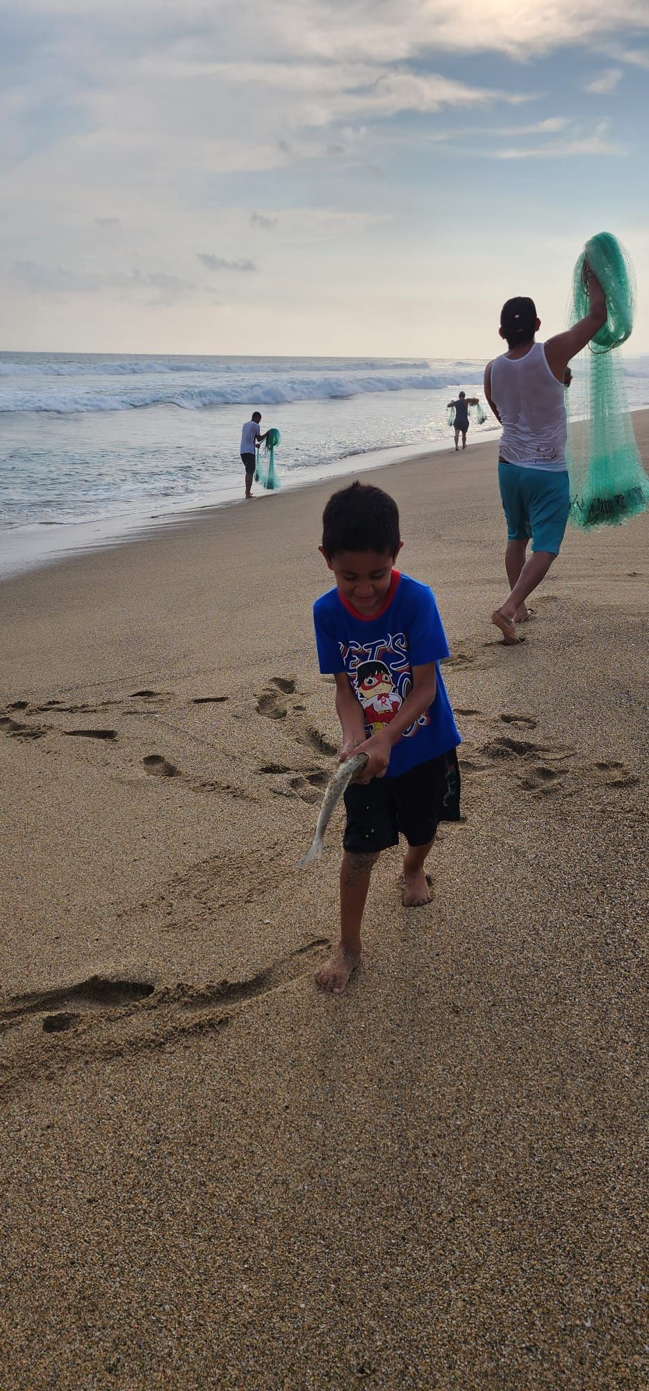 Child running on a pebbly beach while a woman holds a colorful kite near the shoreline.