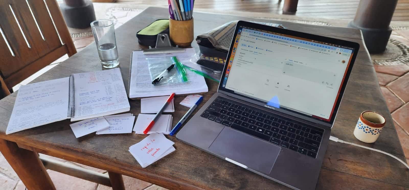 Wooden table with open laptop, scattered papers, pens, and a coffee cup by a window