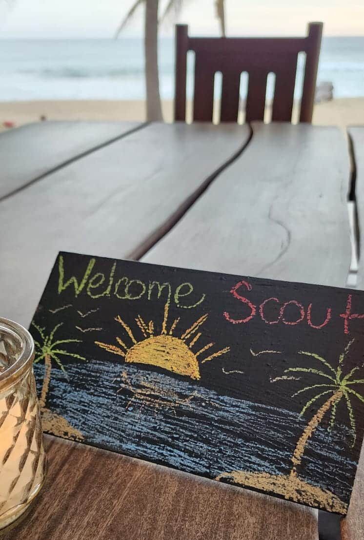 Chalkboard sign saying “Welcome Scout” on a beachside table with ocean in the background
