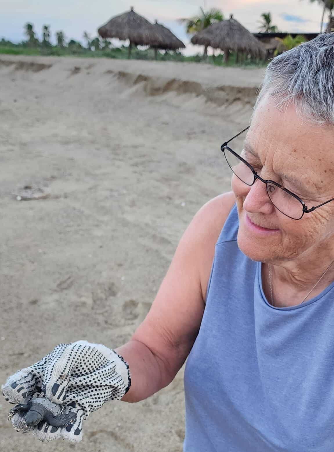 Person in a blue tank top holds a small shell on a beach, with thatched huts in the background