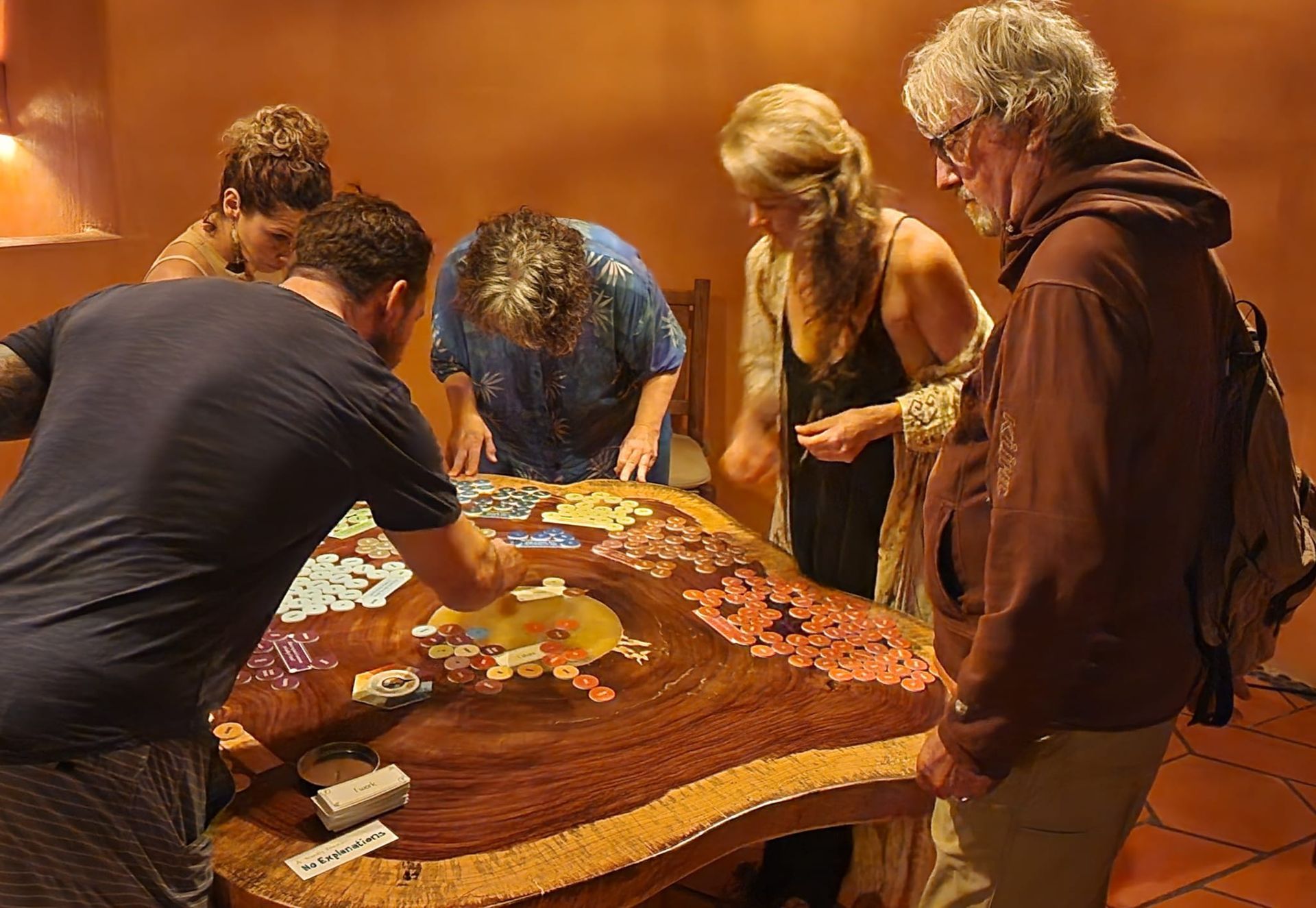 Five people lean over a colorful board game on a table in a warm indoor room.