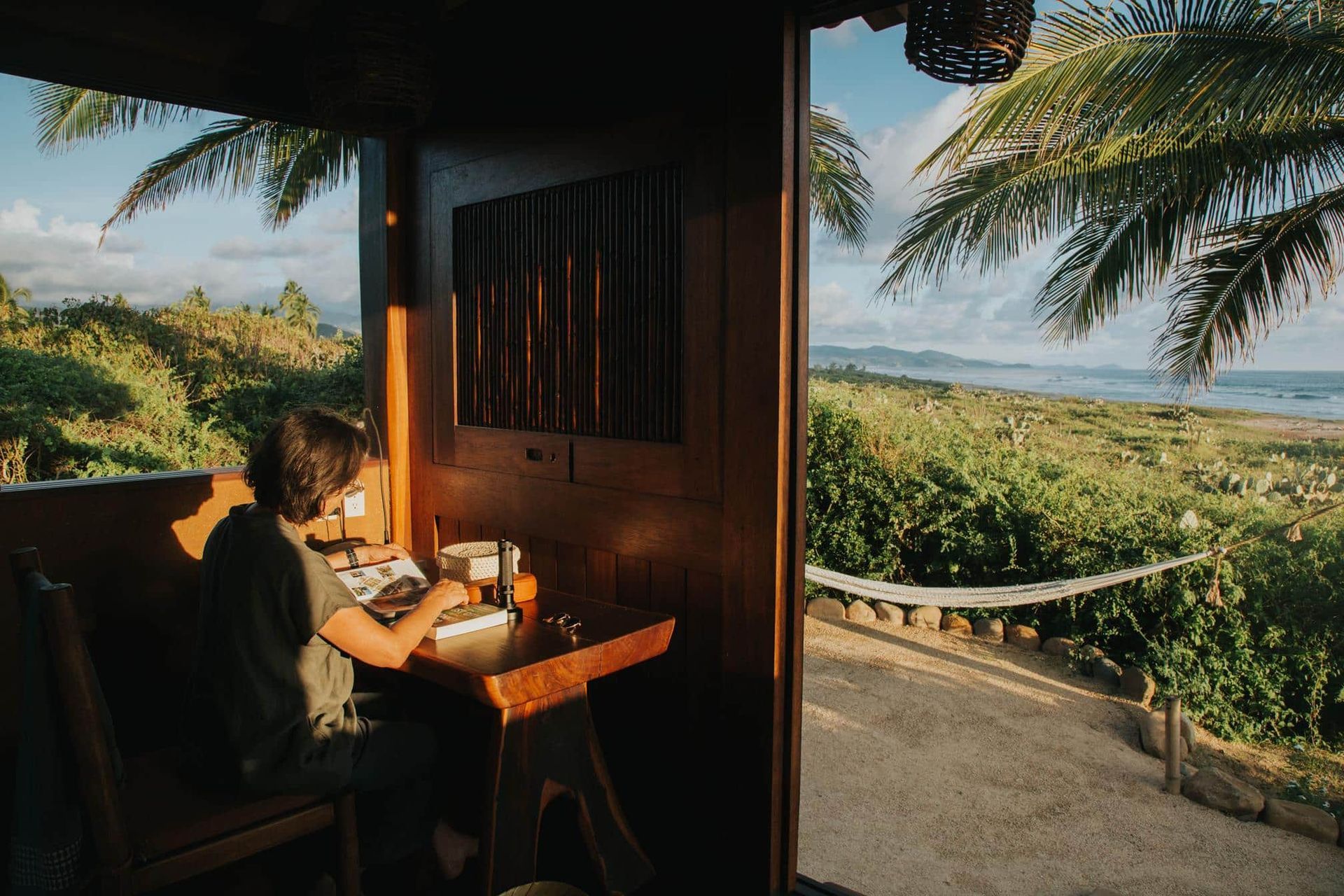 Person writing at a table on a shaded porch overlooking a sunny tropical landscape