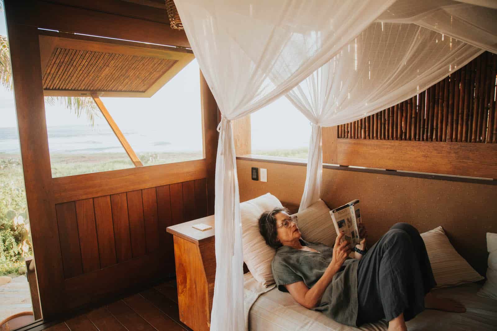 Person relaxing on a bed in a sunlit wooden cabin, reading a phone beside sheer curtains and ocean views