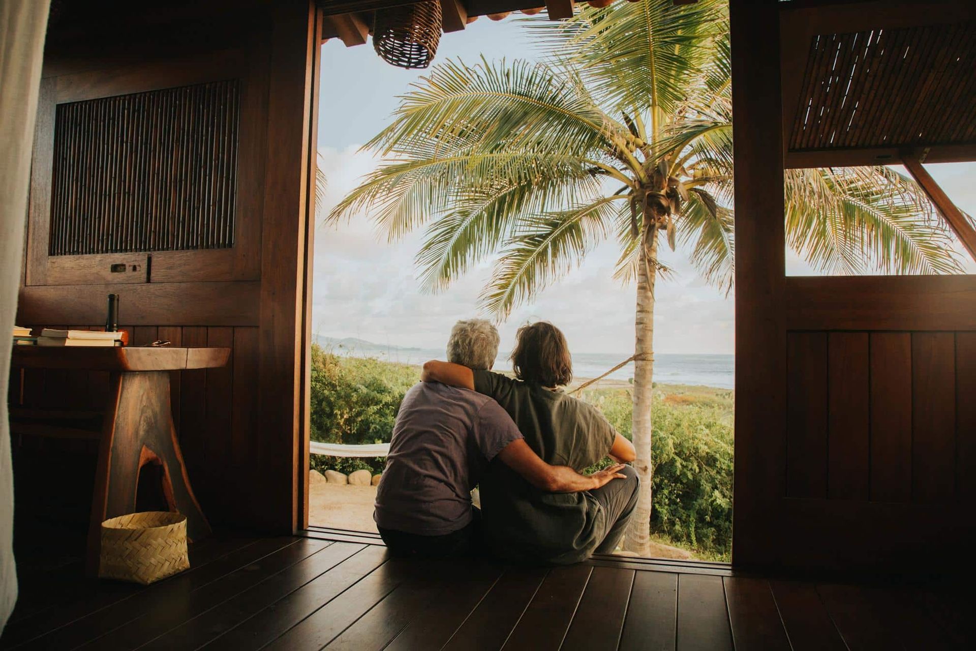 Two people sitting on a porch, looking out at palm trees and a tropical landscape