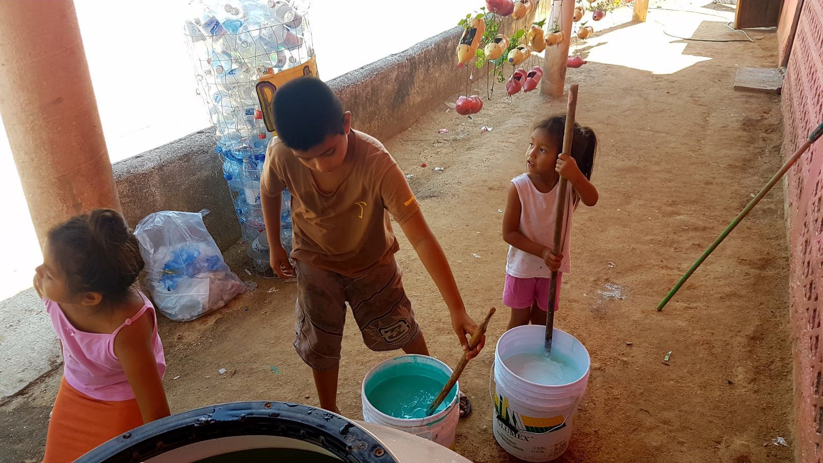 Three children carrying buckets on a sandy floor beside a wall with a painted mural