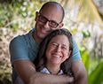 Two smiling people embracing outdoors, with greenery in the background
