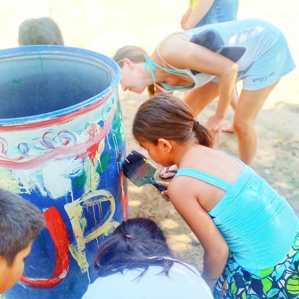 Children painting a large blue barrel outdoors with colorful handprints and designs