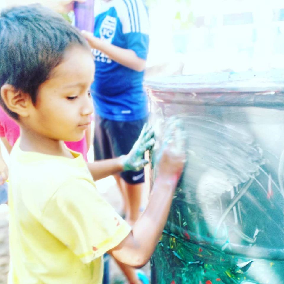 Child painting a large container outdoors with green paint, with another person in the background