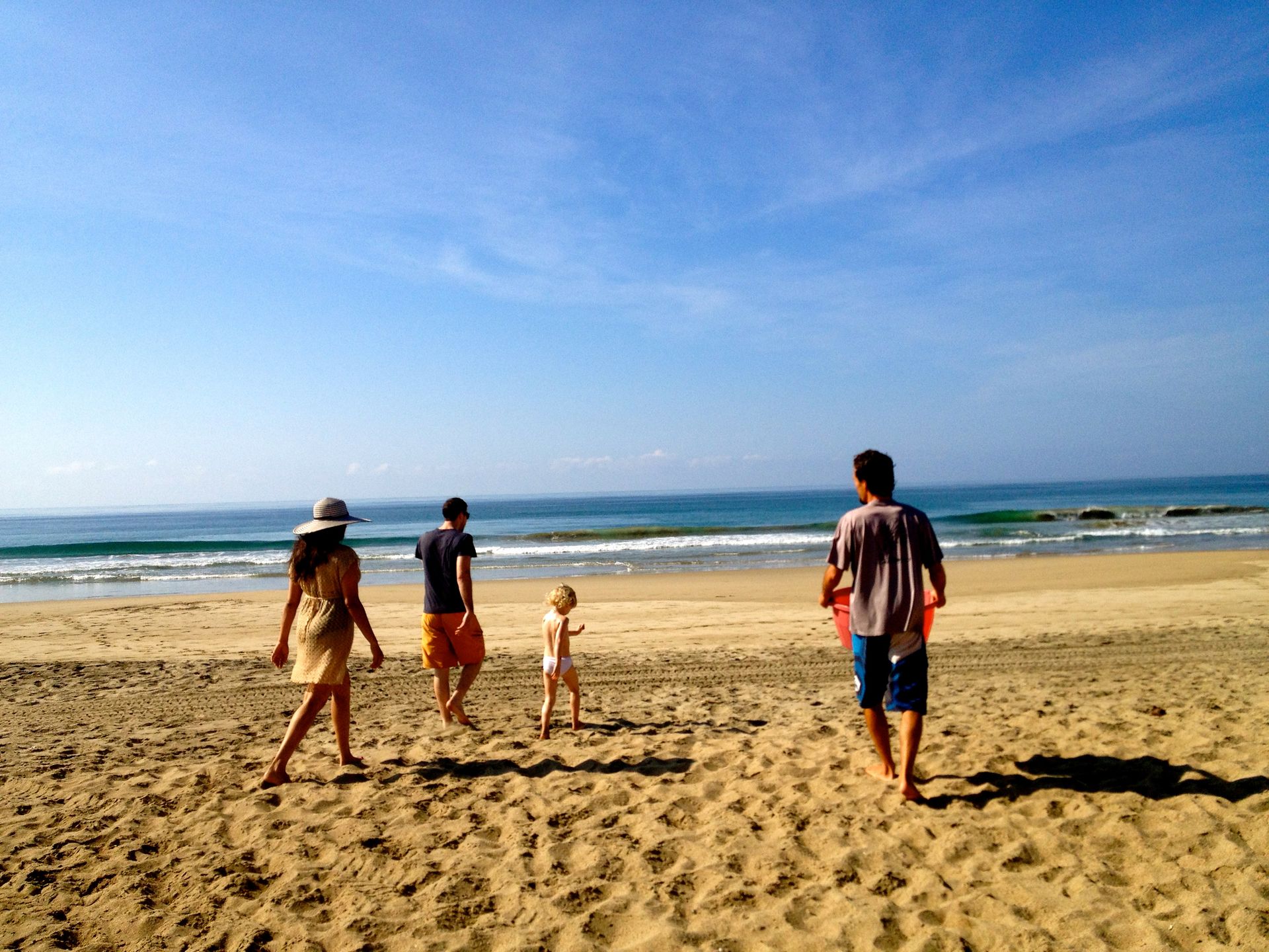 Four people walking on a sandy beach under a blue sky with ocean waves in the background
