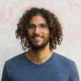 Smiling person with curly hair and glasses wearing a blue sweater against a light background