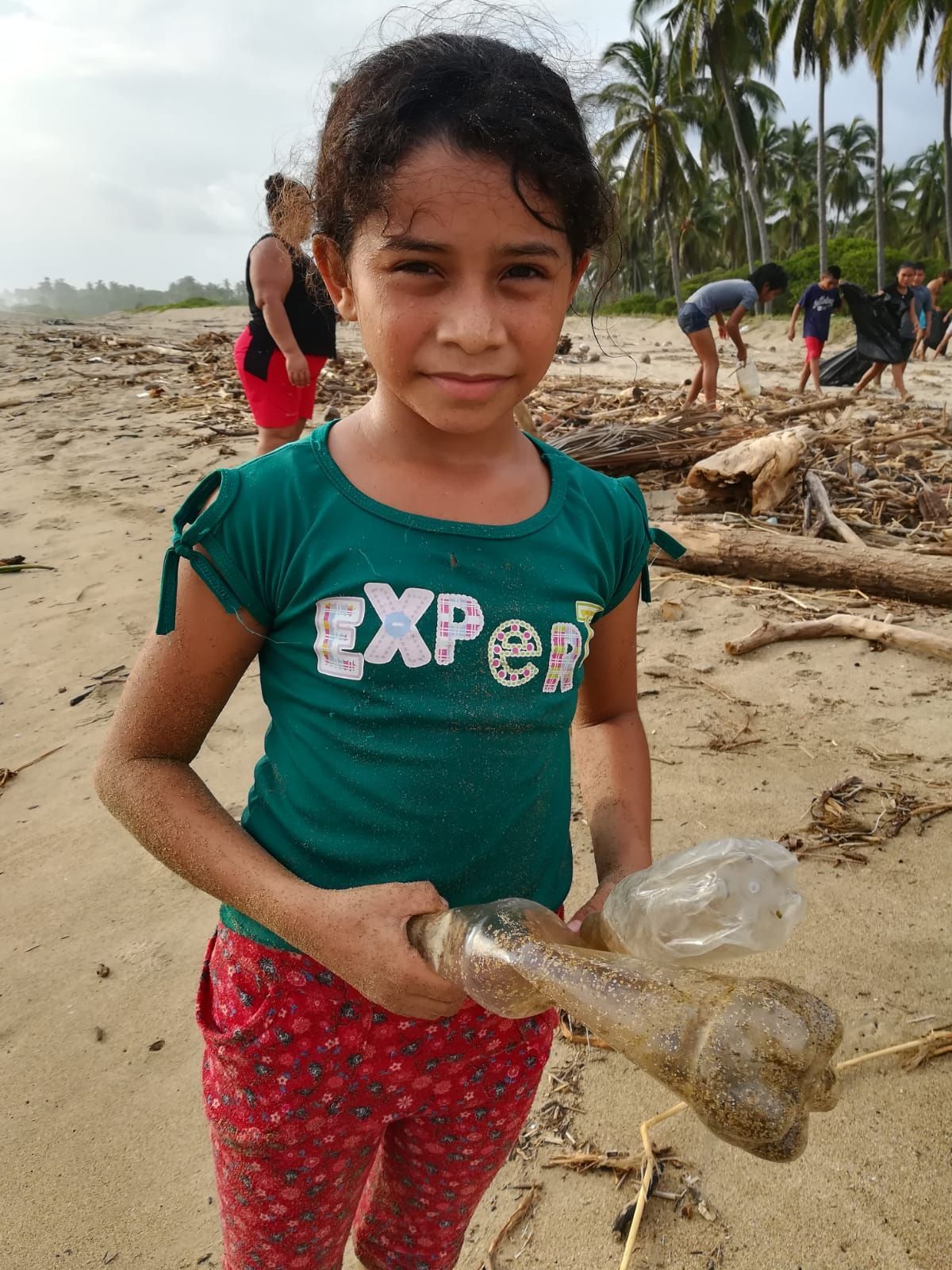 Child on a sandy beach holding a plastic bottle, with people and driftwood in the background.