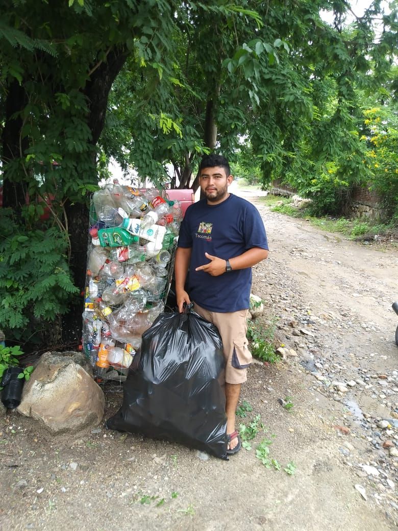 Person standing beside trash bags and a recycling pile on a dirt path under trees