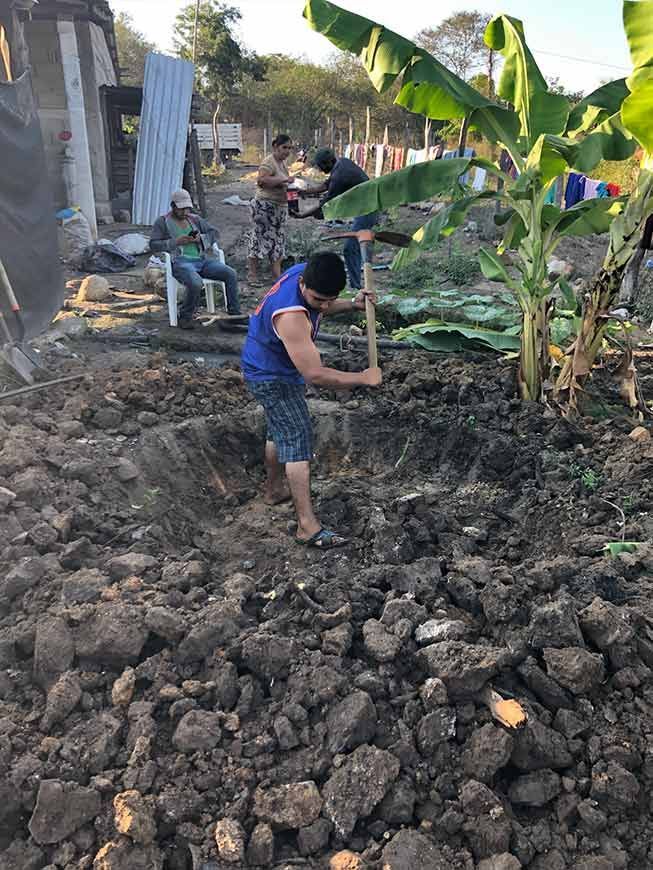 Child digging in a muddy garden plot with banana trees and people working in the background